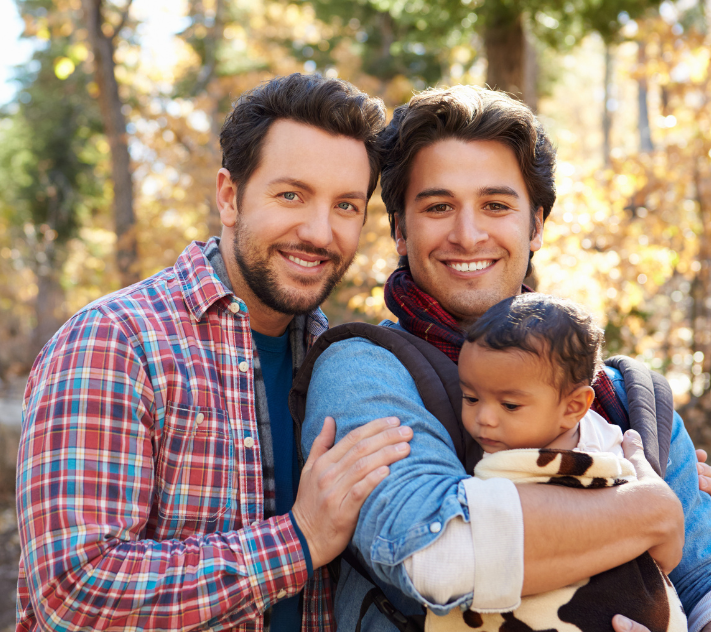 Two fathers holding their son outdoors by trees.