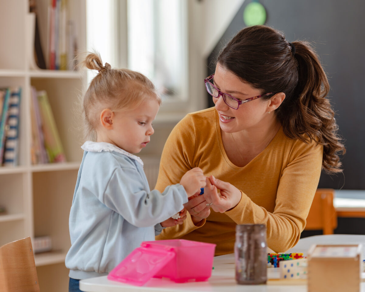 A patient teacher gently guides a shy preschool girl in a classroom activity, helping her adapt to the new environment and build confidence through hands-on learning and engagement