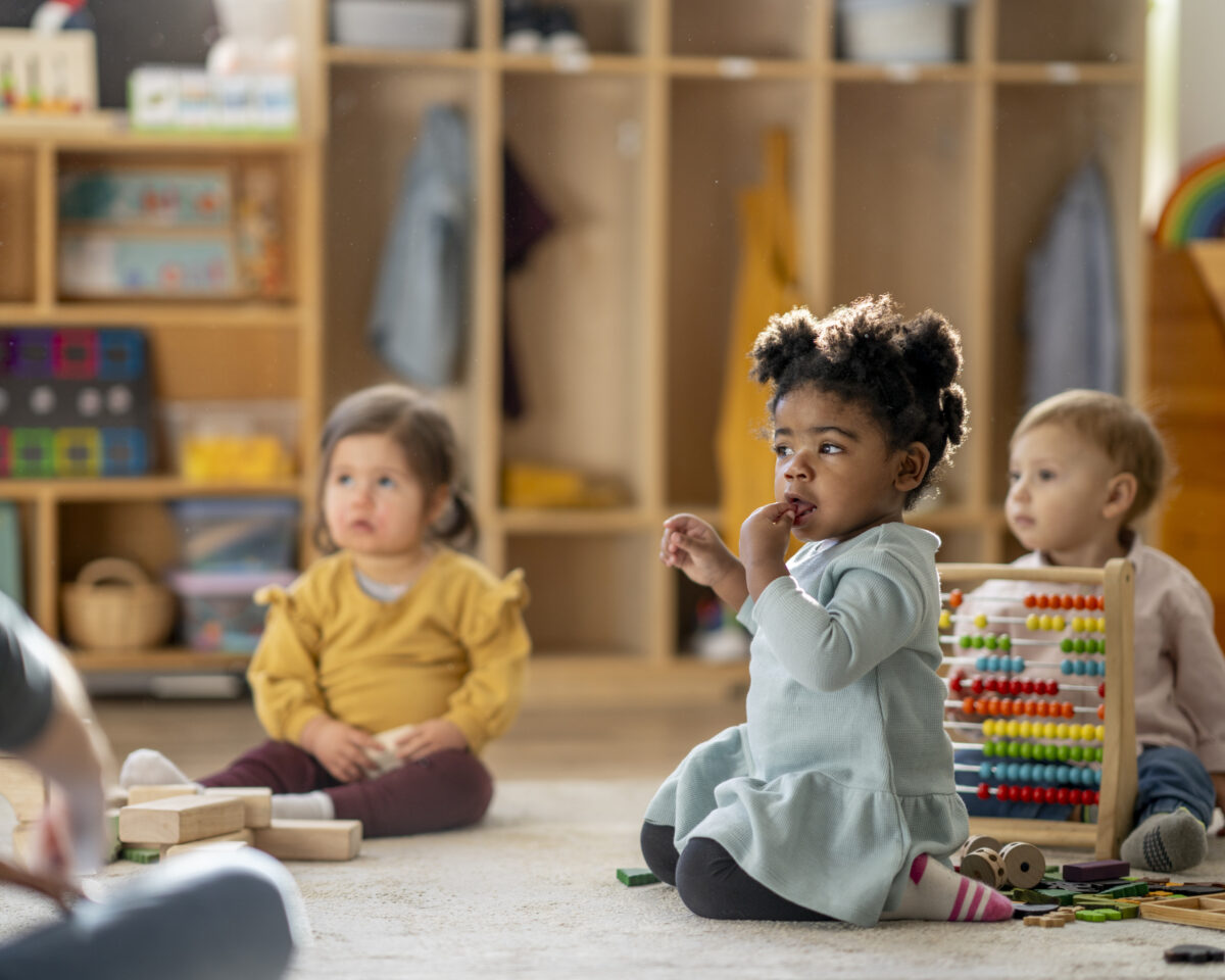 Bright child care scene features diverse children learning and playing with toys, an abacus, and wooden blocks, capturing early education, friendship, and attentive care in a warm, inclusive indoor setting.